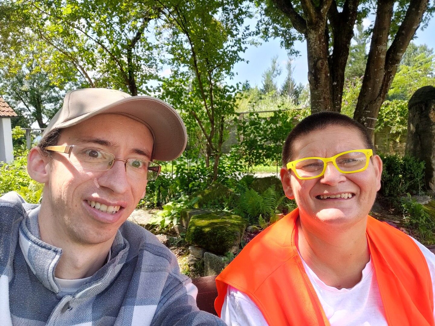 Zwei Personen sind im Selfie. Männlich gelesene Person mit Brille, Kappe, karierter Pullover. Eine weiblich gelesene Person mit gelber Brille, orange Weste, kurze dunkle Haaren. Im Hintergrund Bäume und Natur.